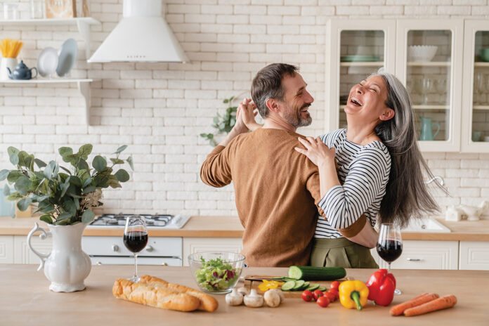 Happy cheerful middle-aged mature couple family parents dancing together in the kitchen, preparing cooking food meal for romantic dinner, spending time together. Active seniors